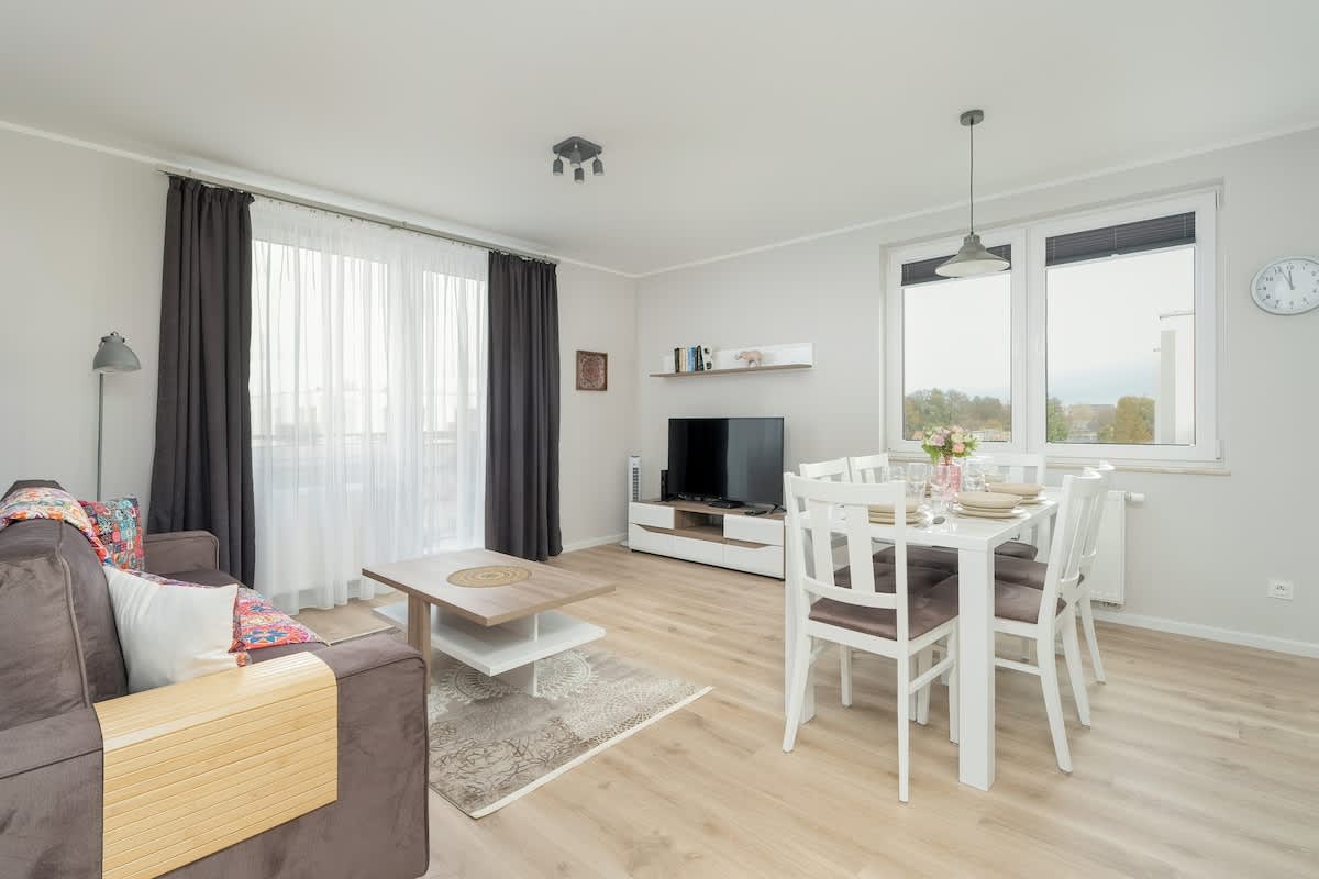 A cozy living and dining area with a light wood floor. There’s a comfortable sofa in the living space and a dining table with white chairs. Large windows bring in natural light.