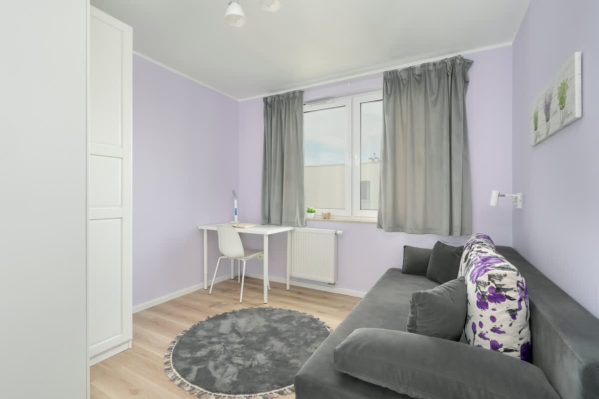 A cozy bedroom with light purple walls and a gray sofa. A small round coffee table and decorative rug add to the relaxed, homey feel.