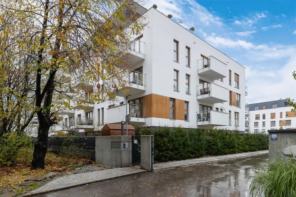 An exterior view of a modern residential building with white and wood-paneled facades, surrounded by well-kept greenery and a paved walkway.

