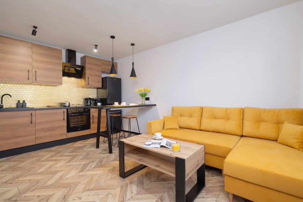 A modern living room with a warm yellow sofa, wooden furniture, and a sleek black kitchen, complemented by stylish lighting.
