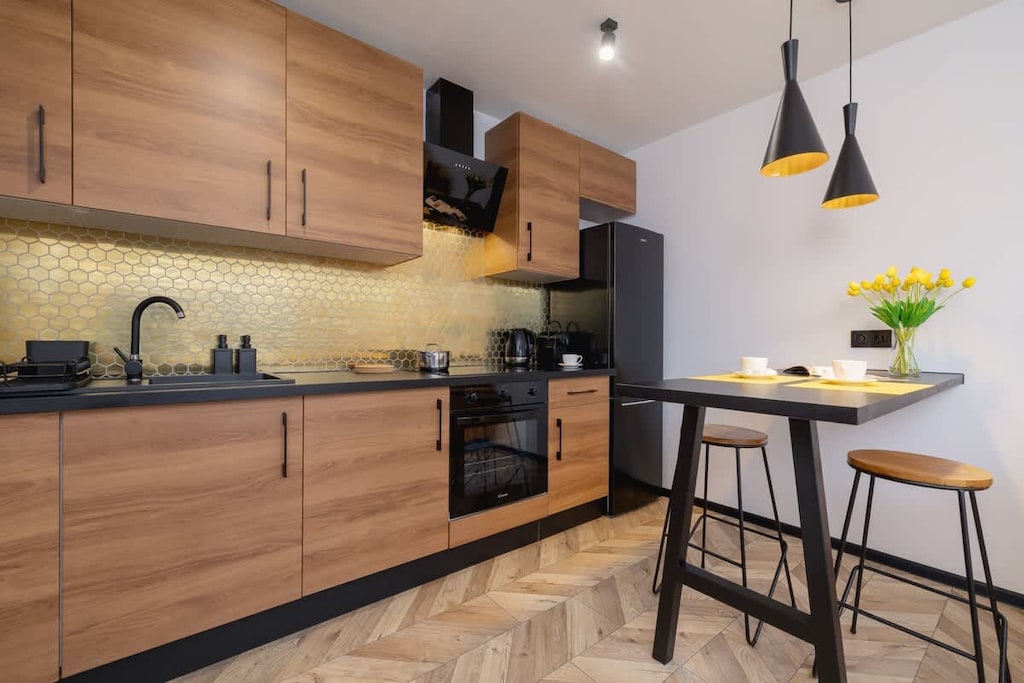 A sleek kitchen with wooden cabinets, a black countertop, and a striking yellow backsplash, paired with black pendant lights and bar stools.
