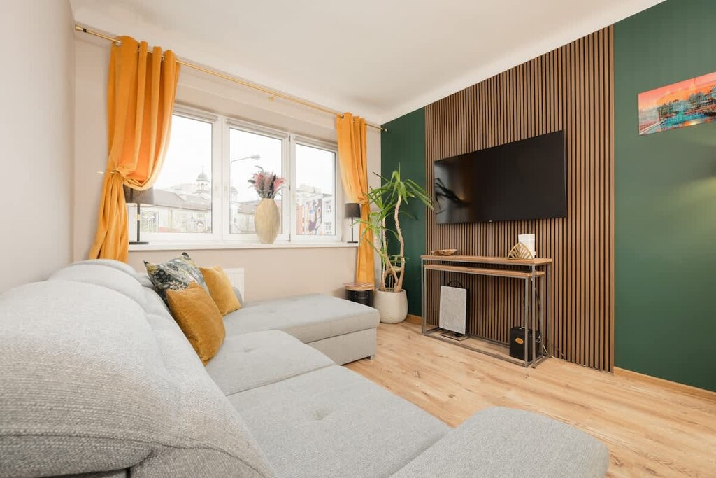 A spacious living room with a large sofa and warm, mustard-colored throw pillows, illuminated by natural light.
