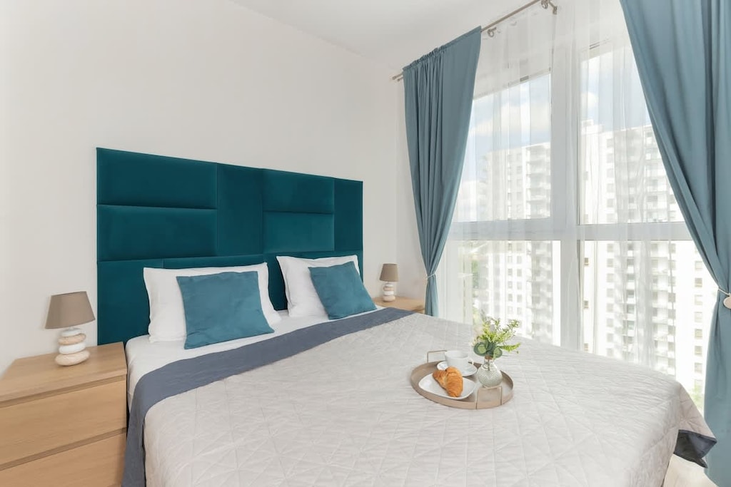 A bedroom featuring a teal upholstered bed, floor-length curtains, and a neutral-toned wall.

