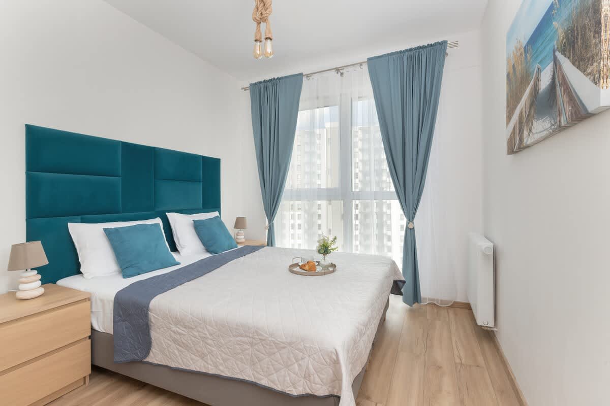 A bright bedroom with a large bed, teal upholstered headboard, and sheer curtains filtering daylight.

