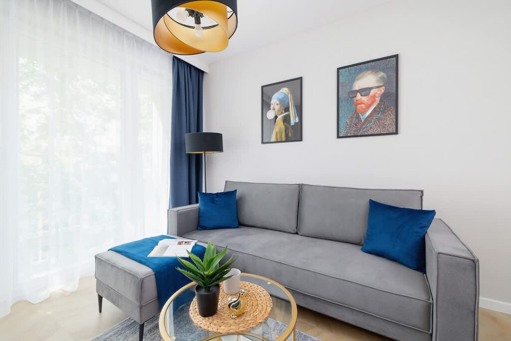 A bright living room with a blue sectional sofa, two framed artworks, and a modern hanging lamp.