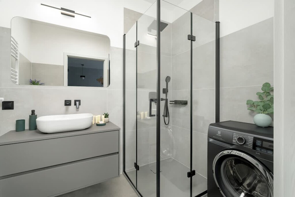 A well-organized, contemporary bathroom with elegant gray tiling, a glass-enclosed shower, and a washer neatly placed under the counter — both practical and stylish.