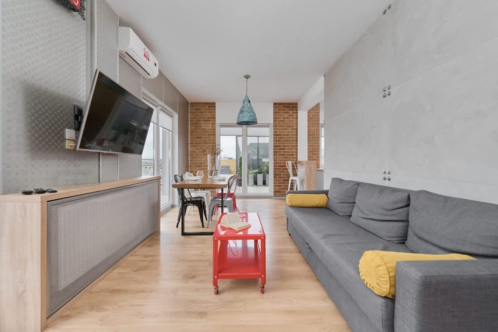 A bright living room with a gray sofa, a red coffee table, and a TV unit, illuminated by natural light from large windows.

