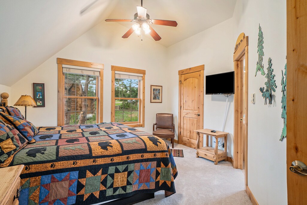 Master bedroom with king size bed, flat screen TV, ceiling fan, vaulted ceiling, and windows with view of outdoors.