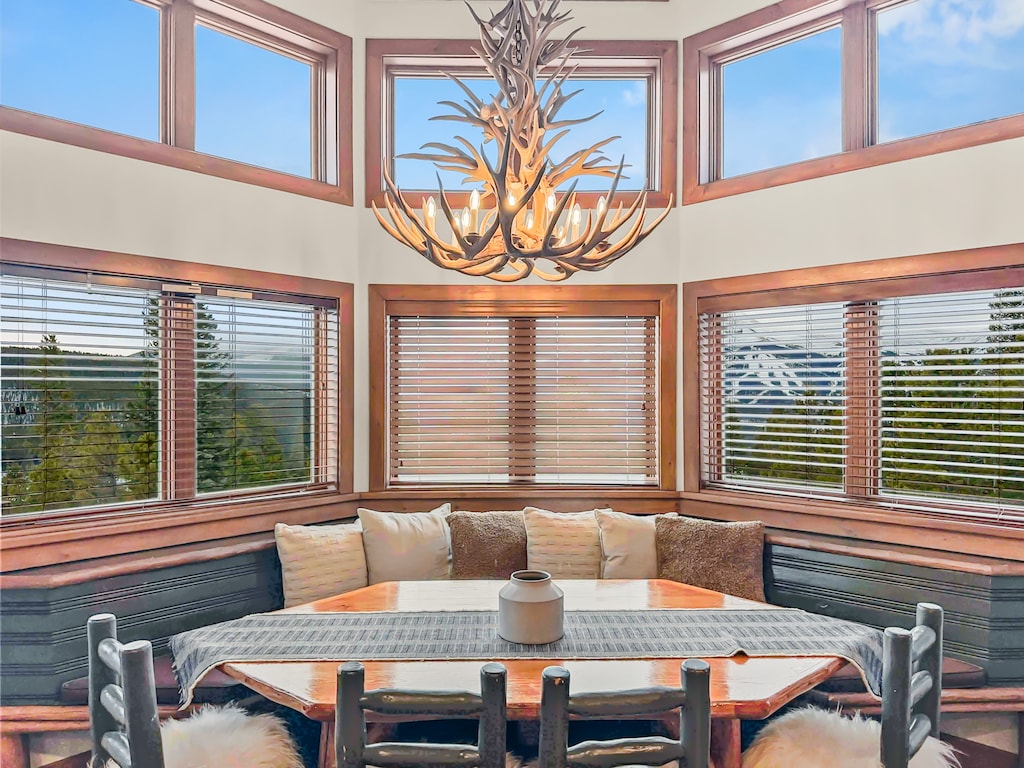 A cozy dining area with a wooden table, cushioned bench seating, and an antler chandelier. Large windows with wooden blinds offer a view of trees and sky.