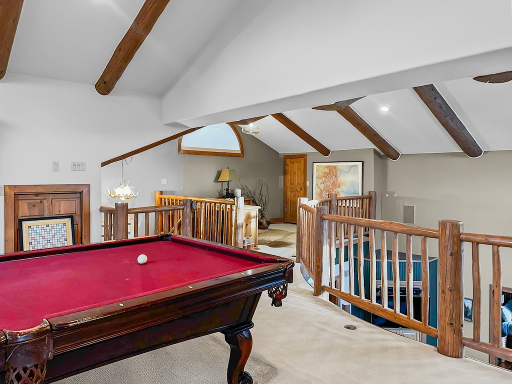 A spacious loft with exposed wooden beams features a red pool table and wooden railings, overlooking a well-lit seating area with artwork and a large rug on the floor.