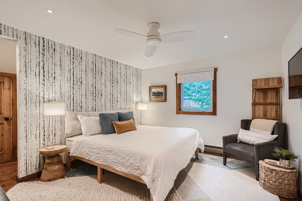A bedroom with a king-sized bed, bedside tables with lamps, an armchair, a window, and a wall-mounted TV. The room features a nature-themed accent wall and a ceiling fan.