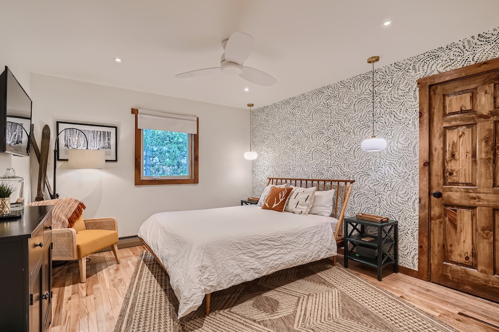 A bedroom with a white quilted bed, wooden headboard, patterned accent wall, wooden door, hanging pendant lights, and a window with a nature view. The room has light wood flooring and rug.