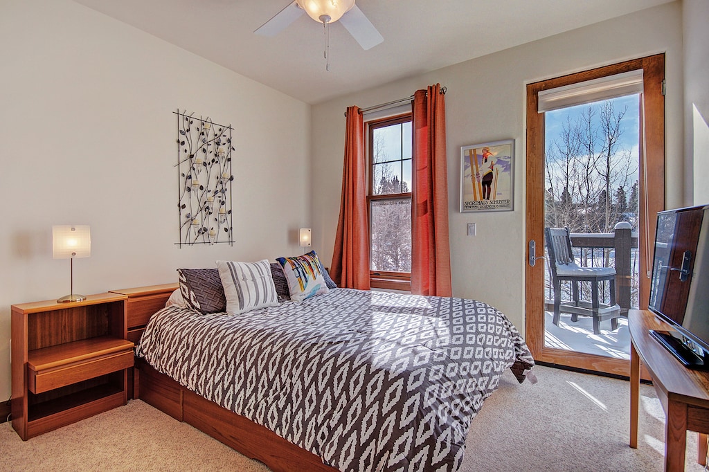 A cozy bedroom with a double bed, patterned bedspread, bedside tables with lamps, two windows with orange curtains, framed art, and a door leading to an outdoor deck with snowy landscape views.