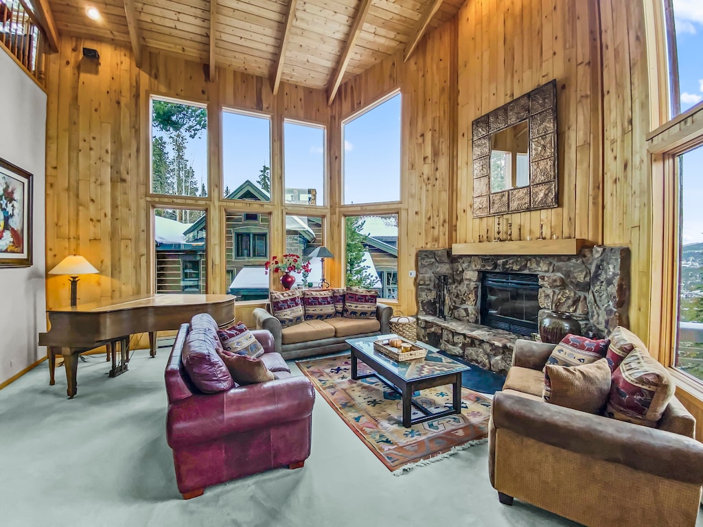 Spacious living room with large windows, wooden walls, vaulted ceiling, stone fireplace, sofas, a piano, glass coffee table, and a patterned area rug. Snowy landscape visible through the windows.