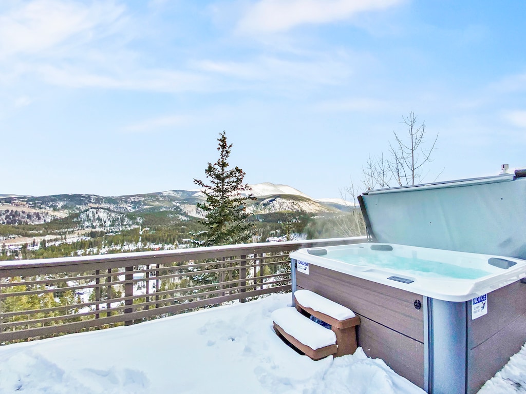 An outdoor hot tub with open lid sits on a snowy deck with a view of snow-covered mountains and trees in the background.