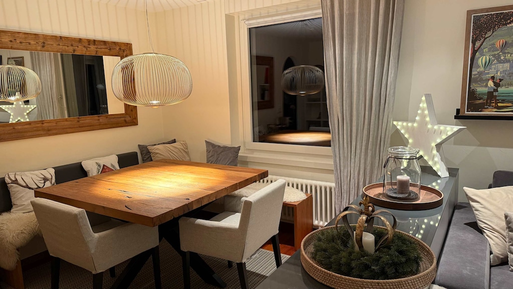 
A dining room features a rustic wooden table surrounded by light-colored chairs and a built-in bench, all lit by a modern, wire-framed pendant light.