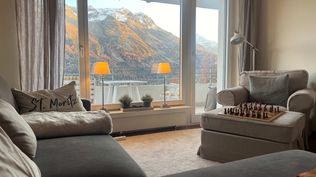 A warm and inviting living room framed by a large window with a breathtaking view of sunlit mountains and a lake.