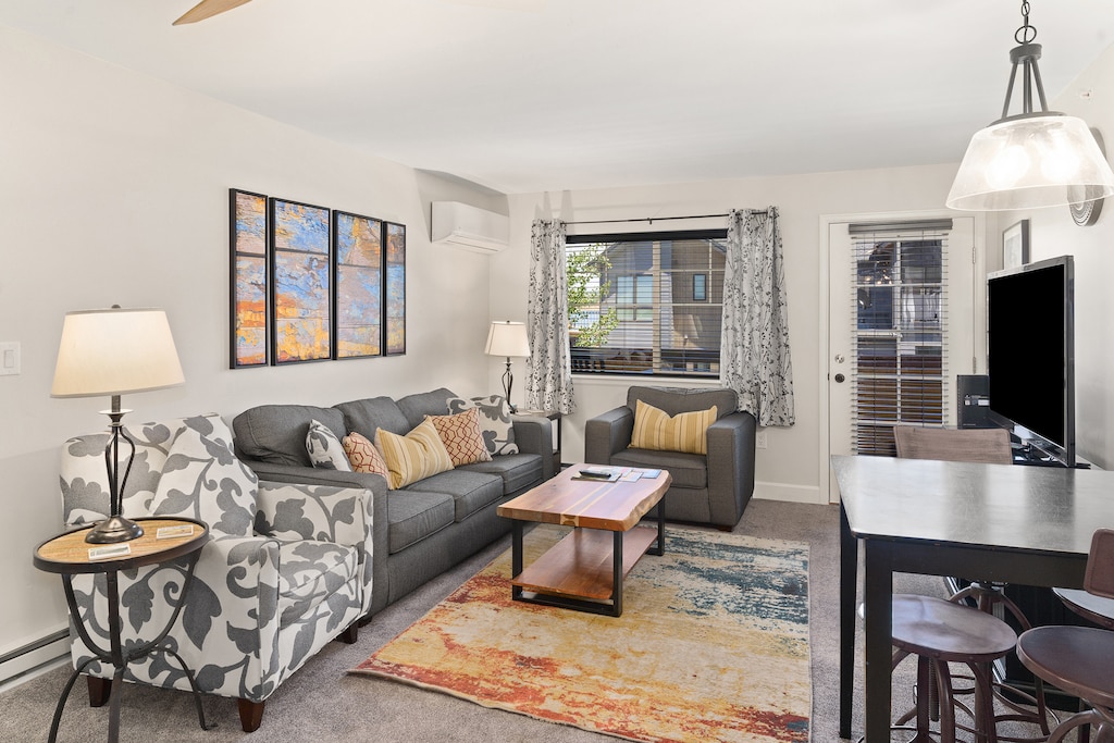 A living room with a grey couch, armchair, wooden coffee table, and rug. There is a TV on a table, a dining table with chairs, artwork on the wall, and a window with curtains.
