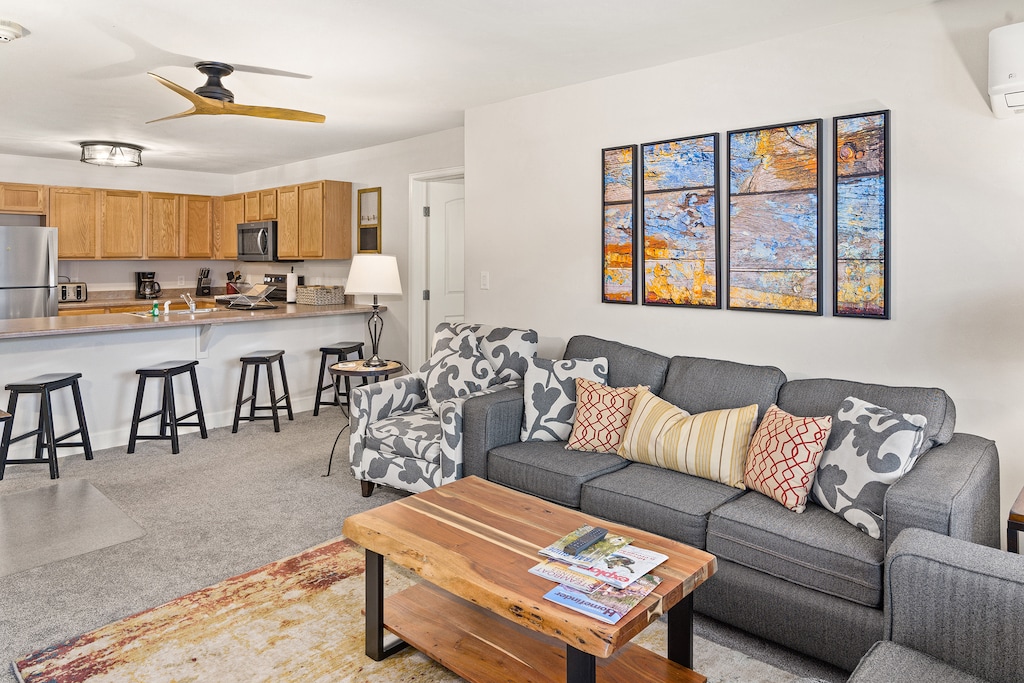 A modern living room with a gray sofa, patterned chairs, a wooden coffee table, and a kitchen in the background. Art pieces are hung on the wall above the sofa, and a ceiling fan is installed.