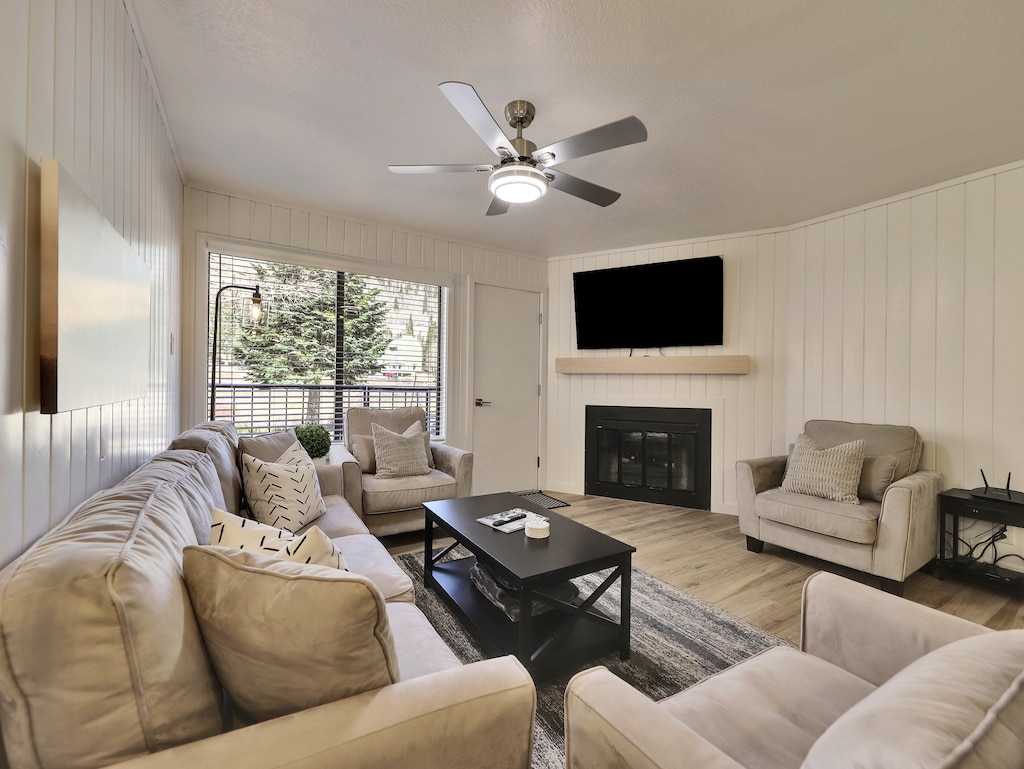 View of the newly updated, light, and airy living room featuring a 58" smart TV, electric fireplace, ceiling fan, and comfortable sofa and armchairs. A glass door leads to the private walk-out balcony