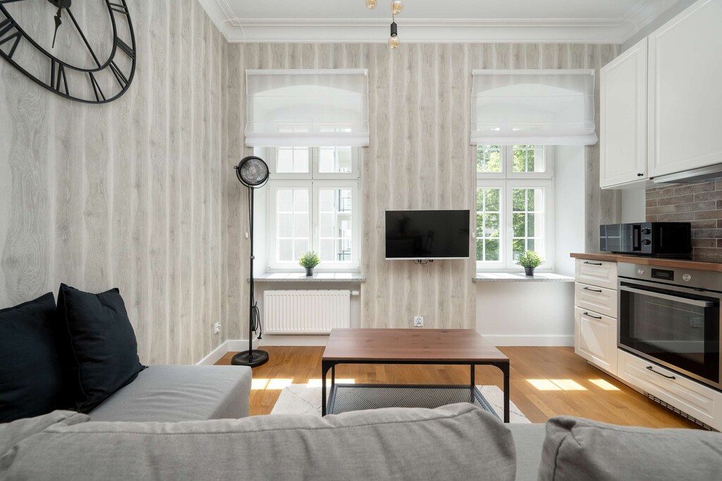 A modern living room corner with a sectional sofa, coffee table, flat-screen TV, and wall clock, all set against patterned wallpaper and large windows.