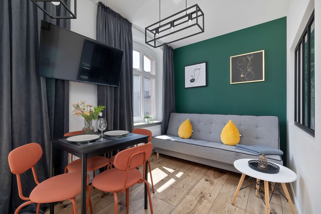 A bright living room with a cozy green wall, orange chairs, and a small coffee table, perfect for relaxing or socializing. Stylish art adds a modern touch to the space.