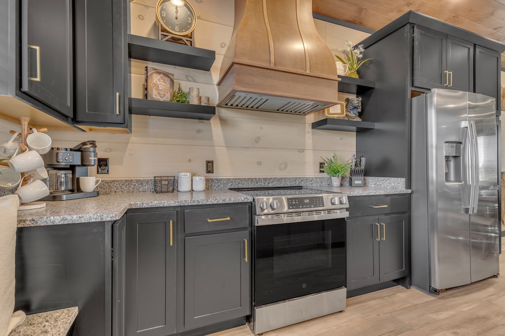 A sleek kitchen with black and gold cabinetry, open shelving, refrigeration, oven and a large range hood.