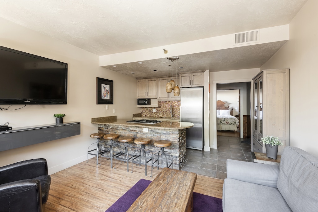 View of the living room with large screen wall mounted TV looking toward the kitchen and bedroom. Hardwood floors in the living room and slate tile floors in the kitchen