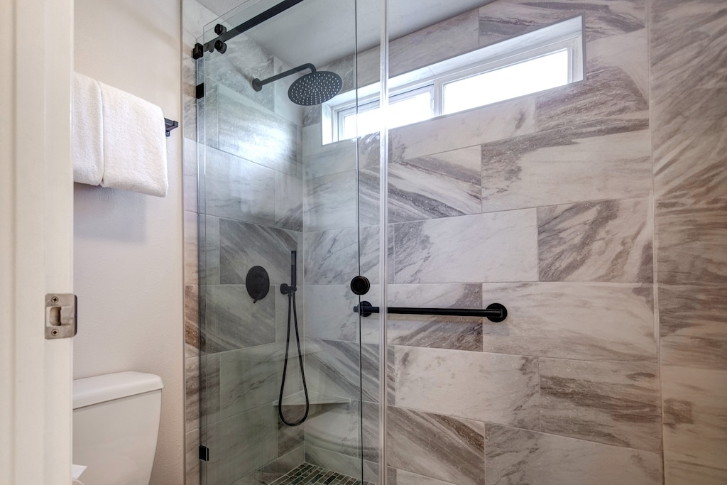 The walk-in shower has grey and white patterned wall tile, black hardware, white toilet, and a white folded hanging towel.
