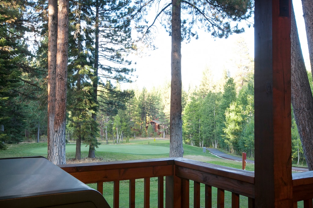 A wooden deck overlooks a lush, green golf course surrounded by tall trees. A grill is partially visible in the foreground.