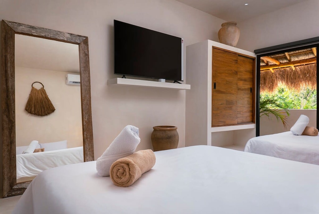 Bright room with minimalist design. It has a TV, AC, closet, and a full-length mirror. Bohemian decorations complete the space.