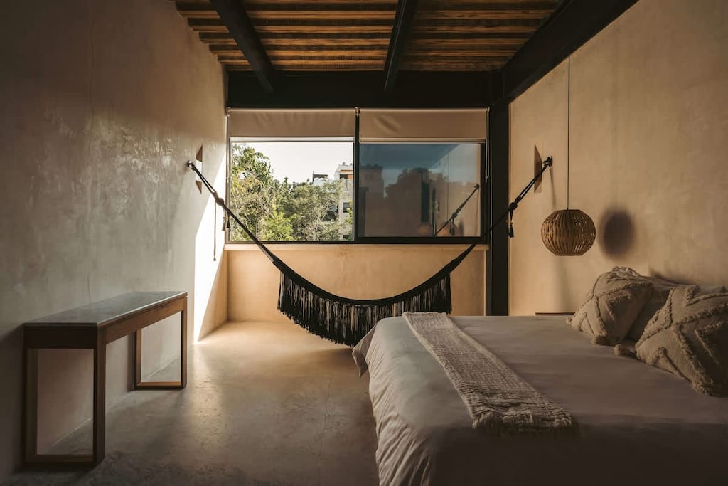 Luxurious and spacious room with a king-size bed. It features a hanging hammock, AC, and wooden furniture. Huge windows cover the wall facing outside, providing an incredible view.
