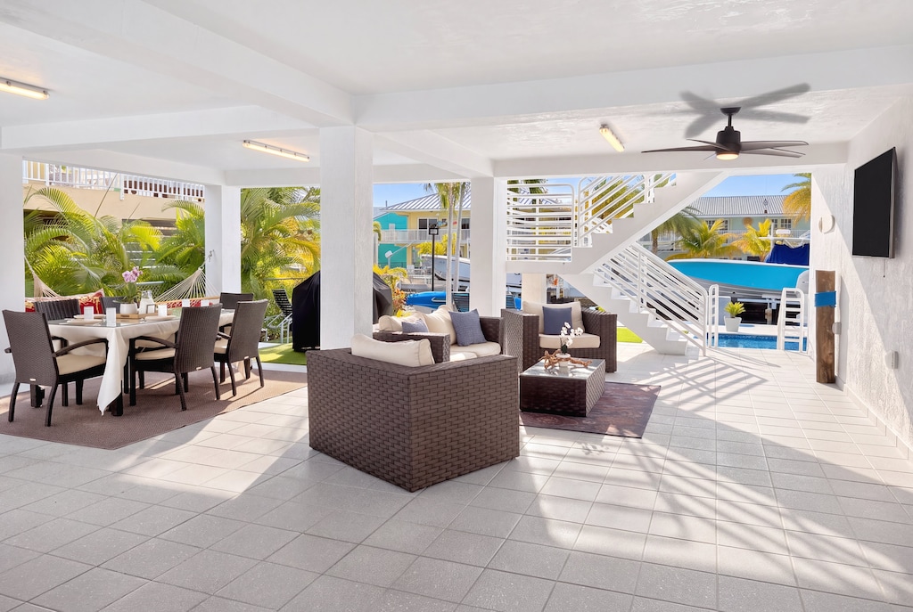 Covered patio | Spacious outdoor living space includes a half bathroom, dining table, couch area, lounge chairs, smart tv, ceiling fan, private boat dock access, water view, and staircase leading to main indoor living area.