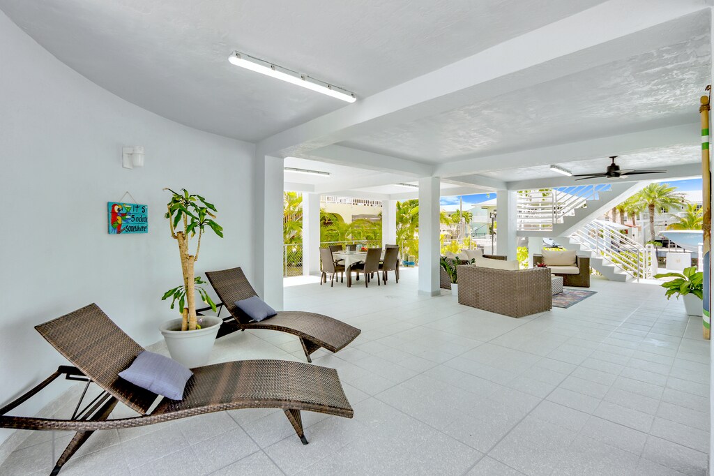 Covered patio | Spacious outdoor living space includes a half bathroom, dining table, couch area, lounge chairs, smart tv, ceiling fan, private boat dock access, water view, and staircase leading to main indoor living area.