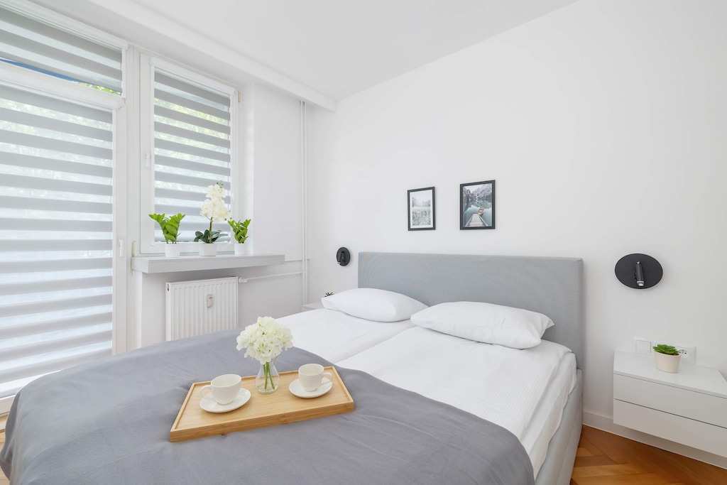 A living space featuring a gray sofa with a breakfast tray set on it and large windows with blinds.