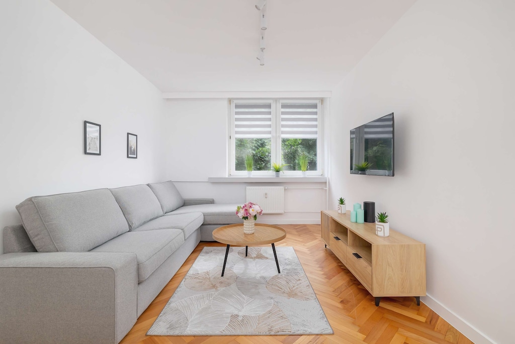 A cozy living room with a gray corner sofa, TV mounted on the wall, white furniture, and a window letting in daylight.