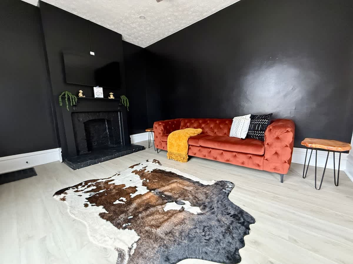 "& Unwind in our chic Jungle House living room in Columbus! Our plush burnt orange sofa with a cozy yellow throw blanket and decorative pillows create the perfect space to relax. The bold black fireplace and flat-screen TV provide a stylish backdrop 