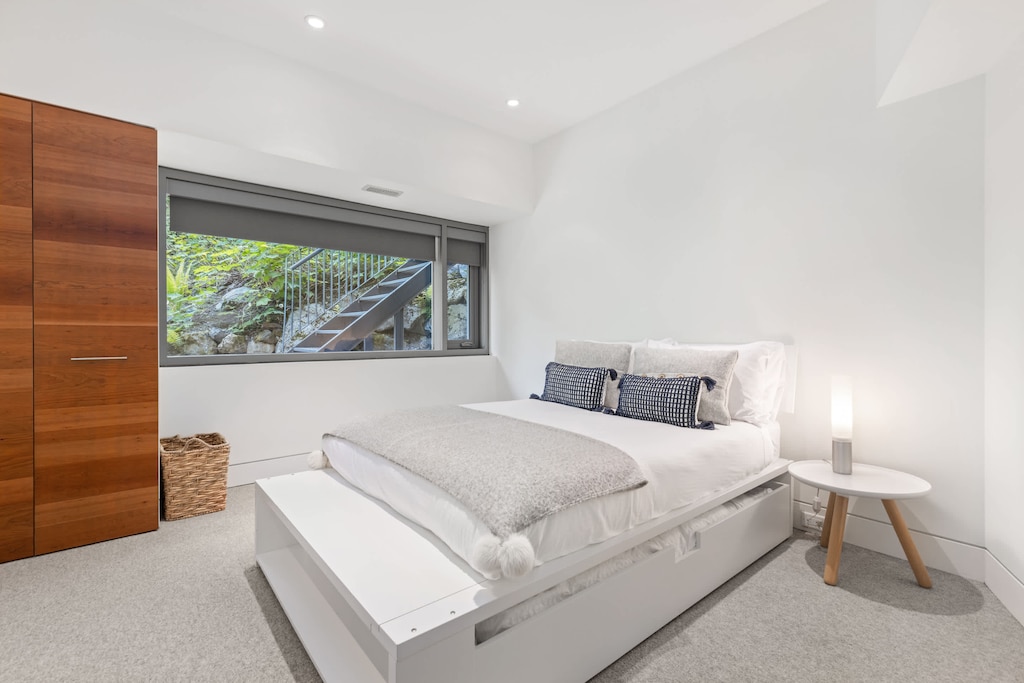 - Peaceful bedroom with soft white tones and wood accents  
- Large window framing lush greenery outside  
- A cozy space designed for rest and calm nights  
