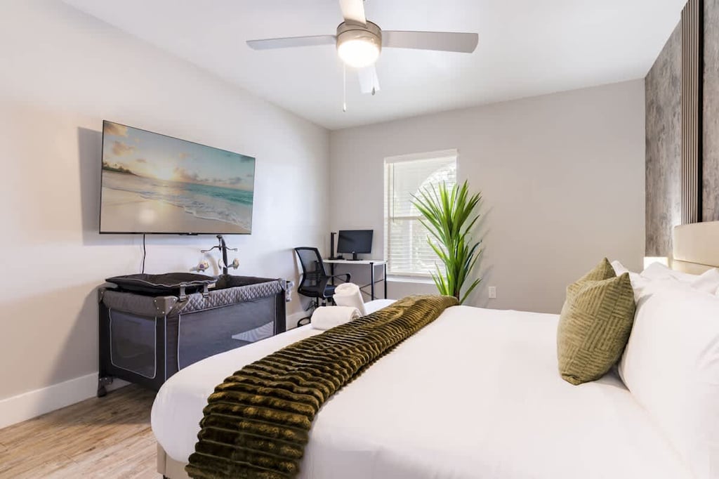 Bright and airy bedroom featuring a king-sized bed, cozy accents, a flat-screen TV and a practical workspace with a crib for family comfort