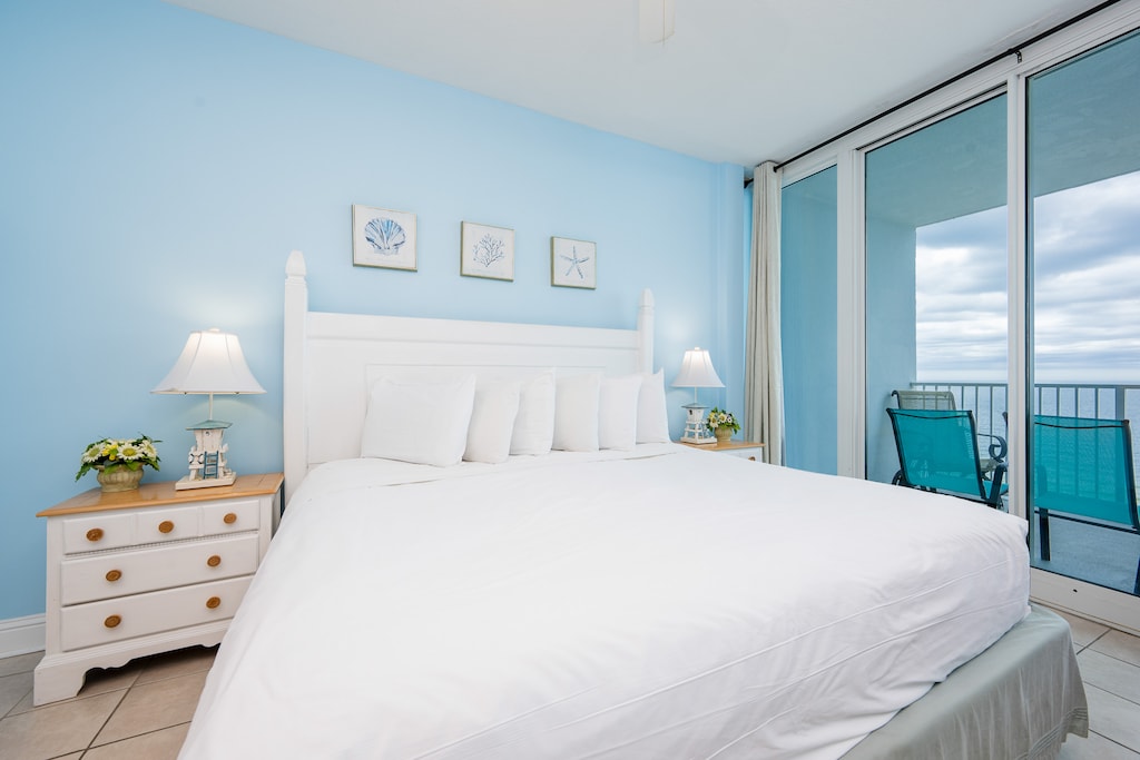 The bedroom features a serene blue wall, a cozy white bed, and a stunning beachfront view with direct balcony access
