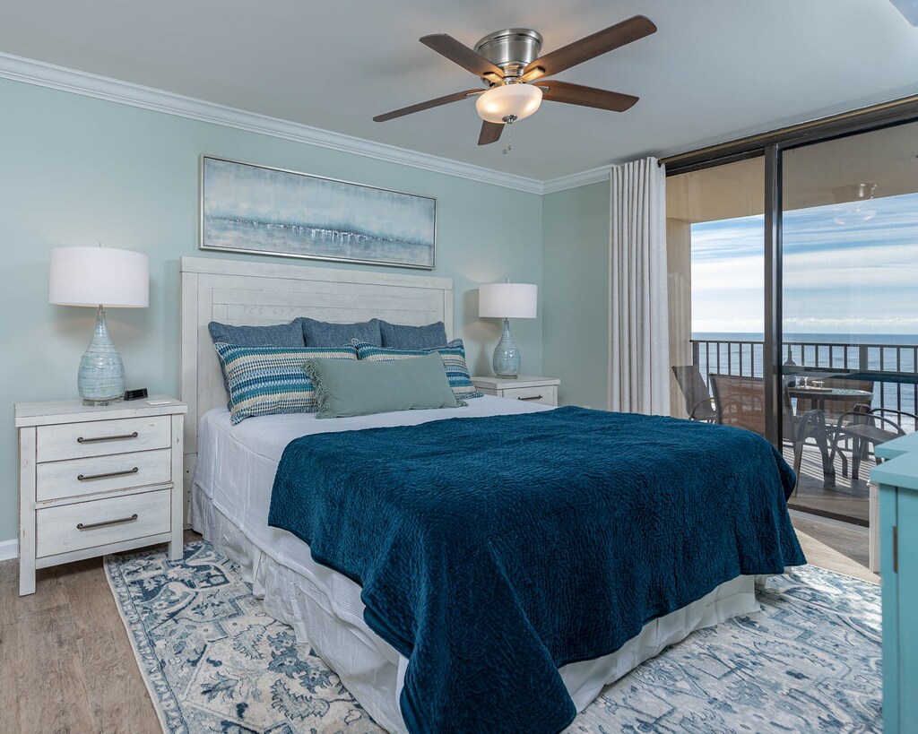 Serene master suite featuring a king bed with a plush teal throw, coastal decor, and a balcony view overlooking the beach