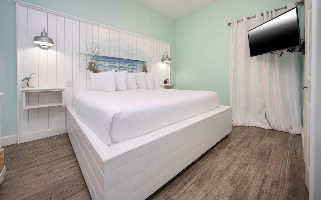 Brightly decorated bedroom features a large white king bed with multiple pillows, coastal artwork, and a mounted TV