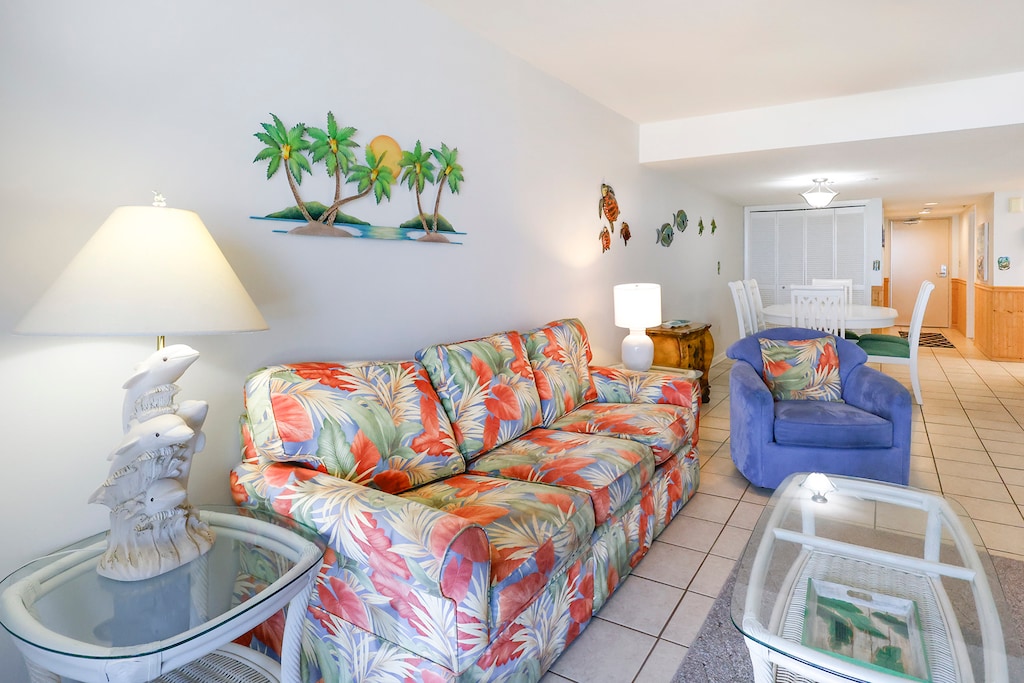 The inviting living space features tropical-themed furniture, a decorative lamp, and cheerful coastal artwork