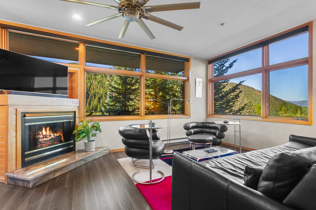 Unwind in this inviting living room where mountain views frame your relaxation. Sink into modern seating by the cozy fireplace, enjoying peaceful forest scenery through expansive windows.