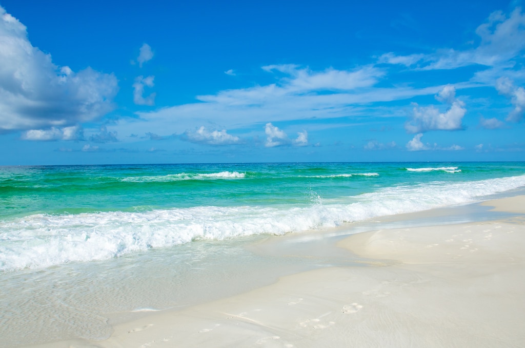 Pristine white sand beach with crystal-clear turquoise waters and gentle waves under a brilliant blue sky with scattered white clouds.