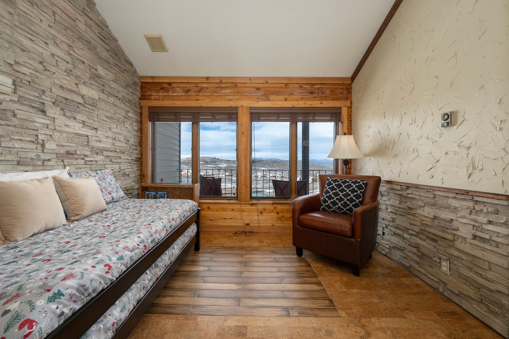 Relax in your cozy bedroom featuring stone accent walls, a comfortable daybed, and panoramic mountain views through large picture windows.