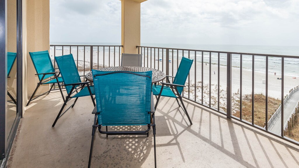 The balcony features a round table surrounded by turquoise chairs, offering a view of the beach and gentle waves below
