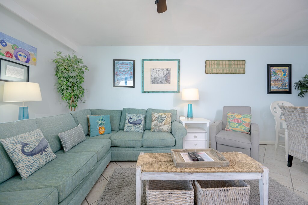 The living room features a light blue sofa adorned with decorative pillows, a woven coffee table, and beach artwork