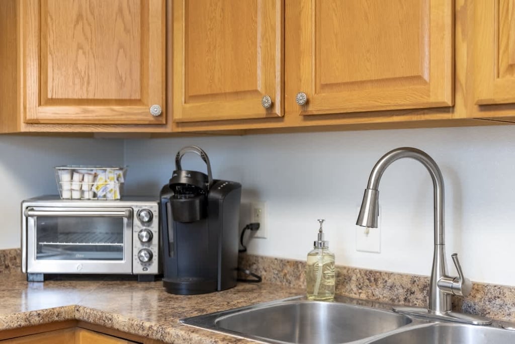 Well-appointed L-shaped kitchen with warm oak cabinets, granite-look countertops, a gas stove, and a double sink under ample cabinet storage. Cozy sign invites you to unwind as you cook and connect. ✨