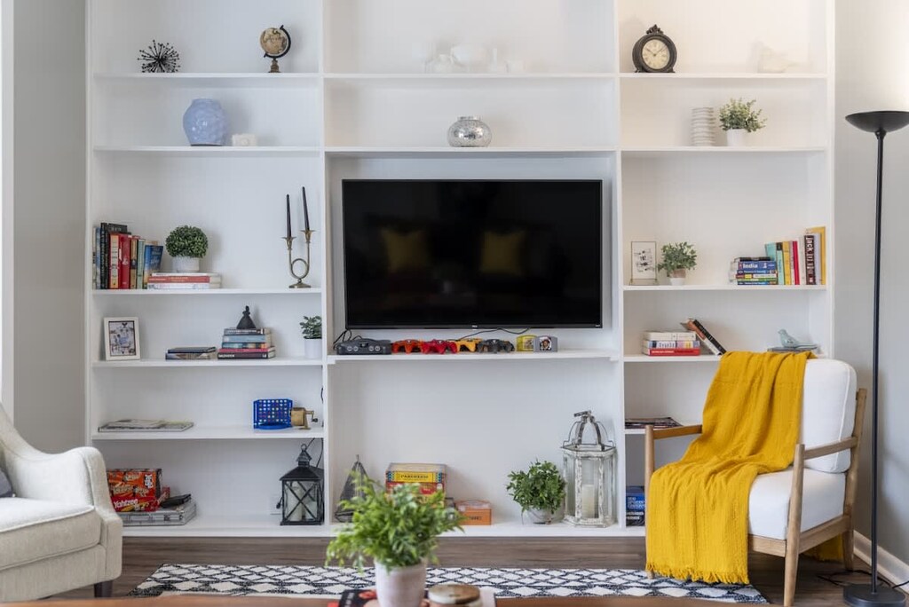 Bright living space with a plush sofa and accent chairs arranged around a live-edge coffee table on a patterned rug. Built-in white shelves flank a wall-mounted TV, while sheer curtains filter soft natural light through large windows. ️